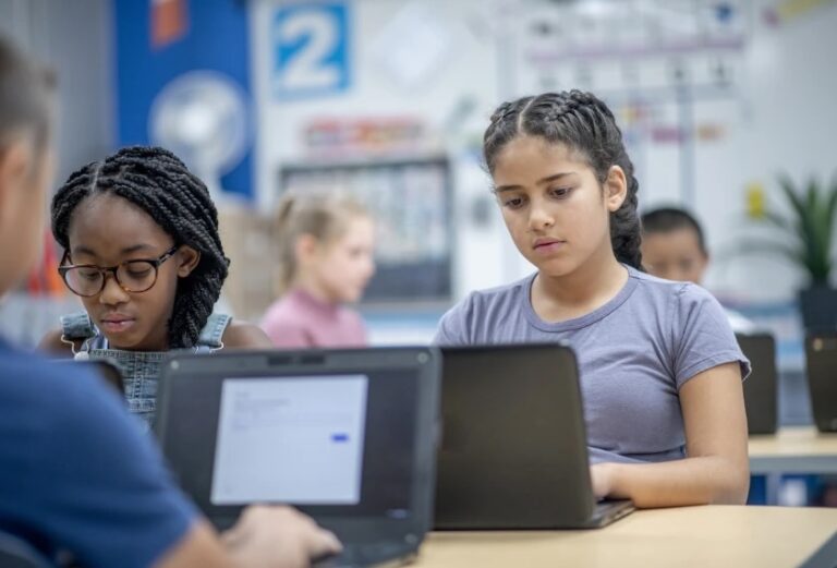 Two young girls working on laptops in a classroom.